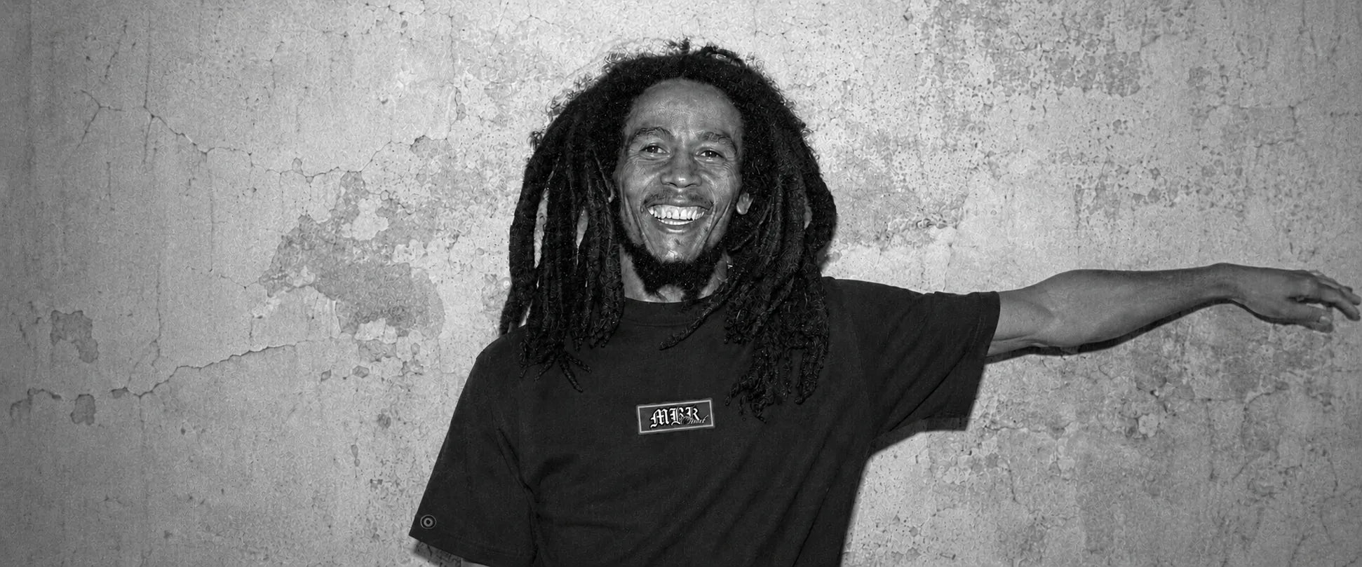 Homem de dreads sorrindo, com camiseta preta MBK, encostado num muro